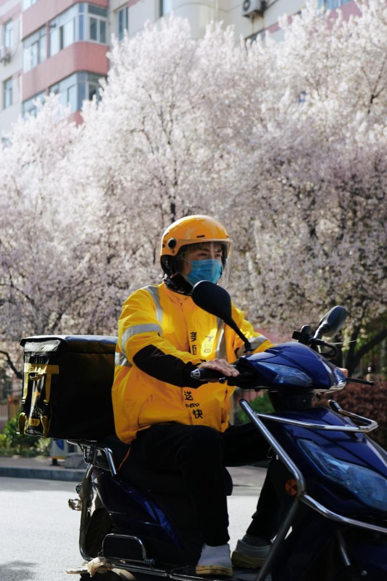 3月12日,佩戴口罩的外卖骑手经过已有樱花开放的街道